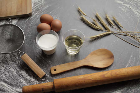 Chicken egg, flour, olive oil, milk, wheat ears, kitchen tool on gray table background. Products for baking bakery products. Cutting board, rolling pin, flour sieve, wooden spoon. For bread or cakeの写真素材