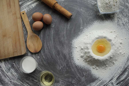 Broken chicken egg in a pile of flour, olive oil, milk, kitchen tool on gray table background. Products for baking bakery products. Cutting board, rolling pin, flour sieve, wooden spoon. bread or cakeの写真素材