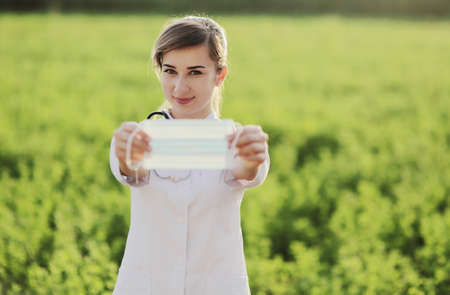 Young woman doctor or nurse showing medical face mask close up. On green grass background. Prevention Covid-19 healthcare concept. Stethoscope over the neck. Female, girl.の写真素材