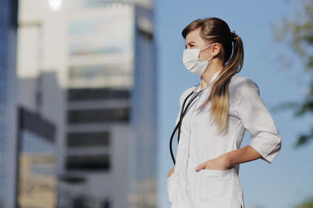 Female doctor, a nurse wearing a protective face mask in the city. Skyscraper, sky. Safety measures against the coronavirus. Prevention Covid-19 healthcare concept. Stethoscope. Woman, girl.の写真素材