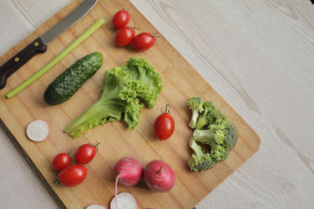 Fresh vegetables on a cutting board on a light wooden table Tomatoes, cucumber, lettuce, broccoli, radish, knifeの写真素材