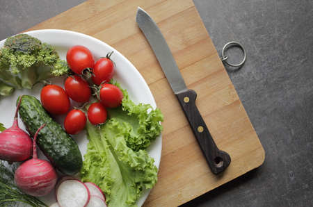 Fresh vegetables in a white plate on a gray table. Tomatoes, cucumber, lettuce, broccoli, radish, cutting boardの写真素材