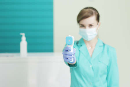Female doctor or nurse in protective mask at hospital holds pyrometer in hand. Safety measures...