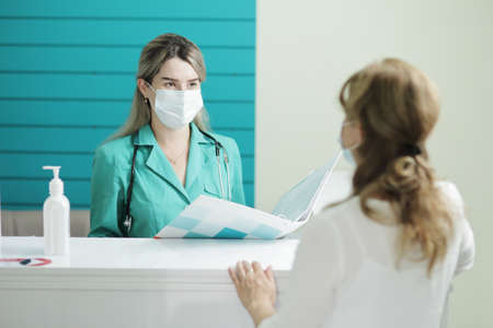 A female doctor or nurse wearing a medical mask talks to a female patient in the waiting room of a hospital. Disinfector on a rackの写真素材