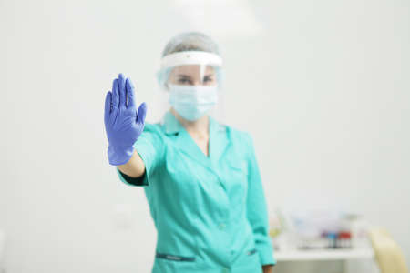 A Female doctor or nurse in uniform, medical mask, protective shield, and gloves shows stop gesture...