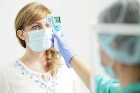 Medical woman in uniform, a medical mask, a face shield, and gloves measures the temperature of a woman with a pyrometer. A female doctor or nurse. Coronavirus COVID-19.の写真素材