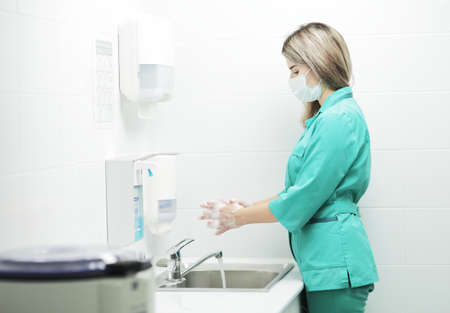 A woman doctor in a protective mask and medical uniform washes her hands thoroughly in the hospital. Soaped hands in foamの写真素材