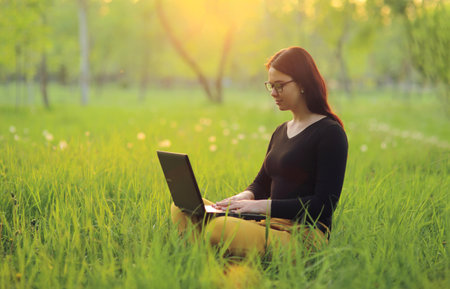 Cute smiling girl with laptop sits on green grass in the park and works remotely as a freelancerの写真素材