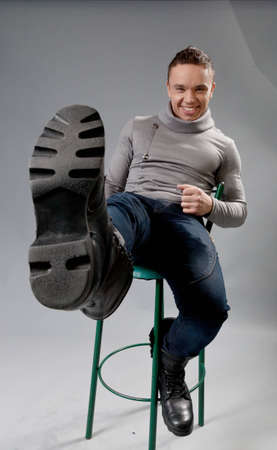 Sexy smiling fit and neat young man in studio playing the fool  Wearing grey turtleneck sweater, dark blue jeans and black boots  Sitting on green bar chair  Fit and neat collection の写真素材