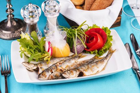 Mediterranean dish, European cuisine. Whole fish grilled capelin, served with a salad of vegetables - greens, arugula, onion rings, tomato, green and yellow pepper and lemonの写真素材
