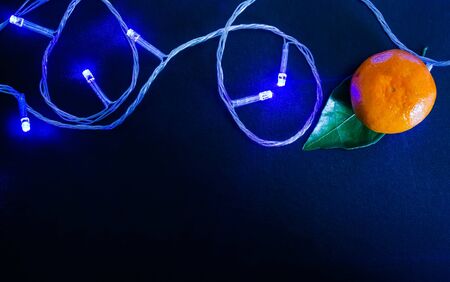 Dark textured Christmas background with tangerines, citrus leaves and a glowing Christmas blue garland. Background for banner, view from above.の写真素材