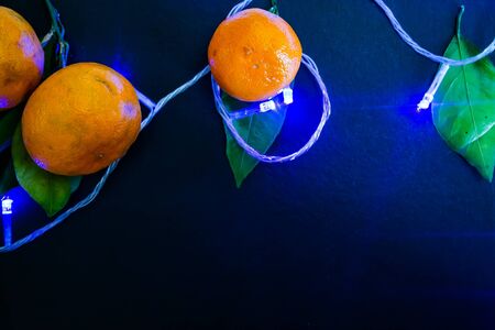 Dark textured Christmas background with tangerines, citrus leaves and a glowing Christmas blue garland. Background for banner, view from above.の写真素材