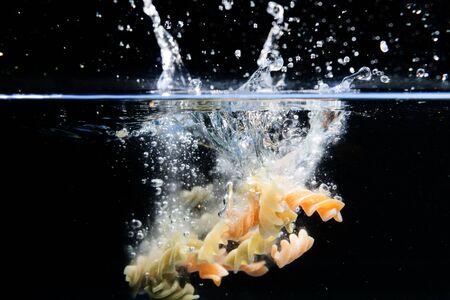 Pasta spirals falling into the water and splashing drops on black background