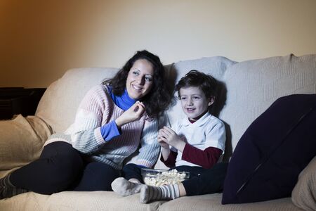 A woman and a child sitting on the sofa, eating popcorn and watching television.の写真素材