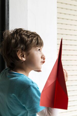 Portrait of a child looking through a window with a red paper plane in his hand.の写真素材