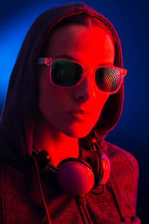 Portrait of a young woman wearing a hoodie, sunglasses and headphones, illuminated with a red light,...