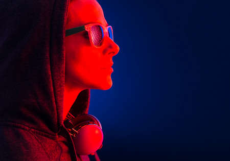 Portrait of a young woman wearing a hooded, sunglasses and headphones, illuminated with a red light,...