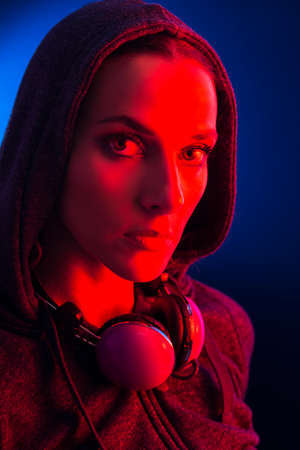 Portrait of a young woman wearing a hooded and headphones, illuminated with a red light on a dark...
