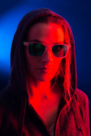 Portrait of a young woman wearing a hooded and sunglasses, illuminated with a red light, on a dark...