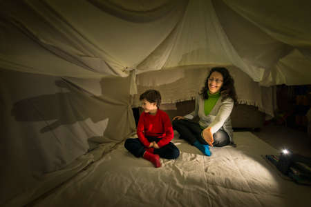 A woman and a child making shadow pictures with the help of a smartphone flashlight in a makeshift tent in their living room. Home lifestyle concept.の写真素材