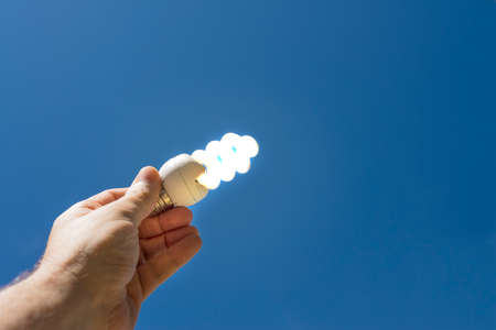 Hand holding a lit energy saving light bulb with a blue sky in the background. Clean energy concept.の写真素材