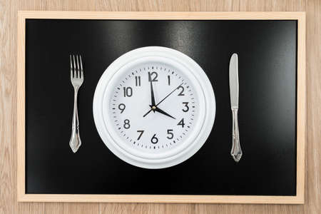 Clock with a knife and fork on a blackboard in the background. Conceptual photograph about intermittent fasting, a diet that brings benefits such as the regeneration mechanism of autophagy.の写真素材