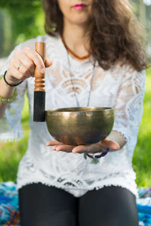 Detail of a woman's hands holding and playing a Tibetan singing bowl during a meditation and music therapy session in a park area.の写真素材