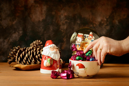 Detail of a hand taking a sweet from a bowl with Christmas motifs that forms part of a still life with Christmas figures, on a wooden table with a dark background.の写真素材