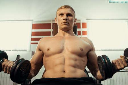 A young athlete with a bare torso doing exercises with dumbbells. Exercises for bitseps.