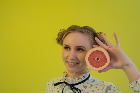 Beautiful girl with funny hair and make-up prof standing with grapefruit in hand on bright yellow backgroundの写真素材