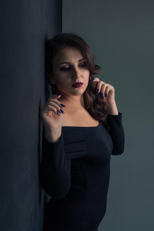 Beautiful girl in a sexy black dress posing while standing against a wall