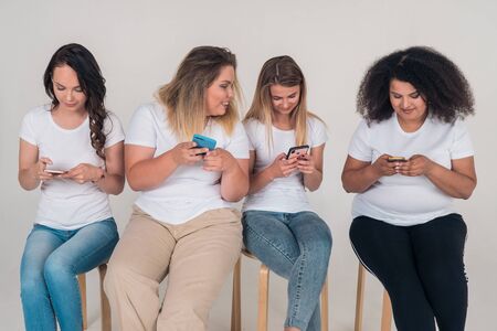 Four young girls use social networks in phones, sitting on chairs on a white background.の写真素材