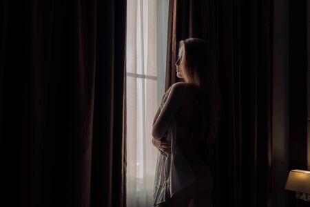A beautiful girl looks out the window covering her body with a mans shirt in the darkの写真素材