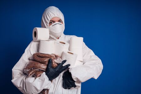 Girl in a protective white suit holds rolls of toilet paper and buckwheat in her hands. Coronovirus panic.の写真素材