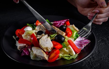 woman hands with knife and fork eat Fresh salad with chicken breast, cheese, black olives,red pepper, lettuce, fresh sald leaves and tomato on a black plate on Dark grey black slate backgroundの写真素材