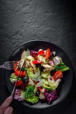 woman hands with knife and fork eat Fresh salad with chicken breast, cheese, black olives,red pepper, lettuce, fresh sald leaves and tomato on a black plate on Dark grey black slate backgroundの写真素材