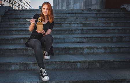 young woman or teenage girl eating asian fast food from takeaway box sitting on the steps on city street. Thai noodles in paper box takeaway street foodの写真素材