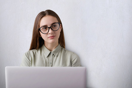 A photo of cute young student with long dark hair wearing eyeglasses working with her laptop at workplace looking away having dreams about something being tired. Educational and studying concept.の写真素材