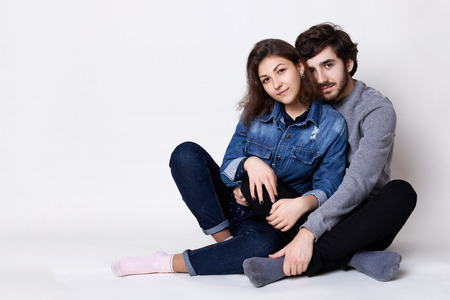 A happy couple sitting crossed legs on the floor. A bearded guy embracing her girlfriend with love. Two people sitting close to each other barefoot isolated over white backgroundの写真素材