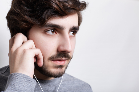 Music and technology concept. A stylish hipster listening to audiobook or radio on cell phone with earphones looking aside touching earphones with his hand isolated over white spaceの写真素材