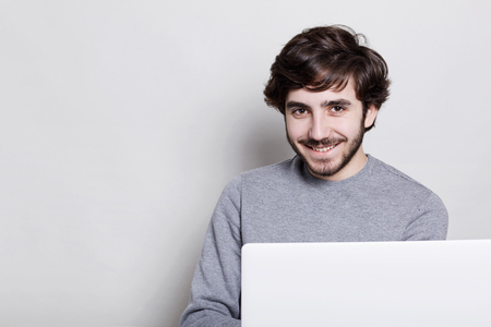 Emotions and feelings. A photo of positive man with cheerful expression having stylish beard and hairstyle dressed in casual clothes sitting in front of his laptop and smiling being pleasedの写真素材