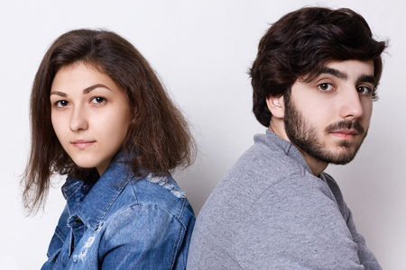 Two young people sitting back to back looking directly into camera. A young couple: brunette girl in jean clothes and stylish bearded man in grey sweater sitting backs relaxingの写真素材