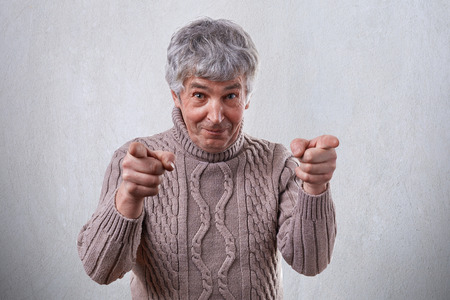A happy mature man with smile on his face standing against white background pointing his index fingers at camera. Senior mature, elderly man pointing at you. Positive human emotions and feelingsの写真素材