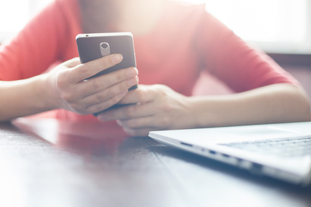 Young woman holding mobile phone and typing messages while sitting at wooden table with open laptop computer. Young woman making online shopping by laptop and smartphone. Modern technology and people.の写真素材