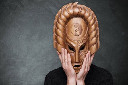 A person wearing wooden mask yin yang symbolizing harmony holding his hands on the mask standing over gray background. Philosophical sense. Dark and light sides of life.の写真素材