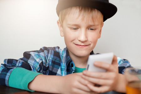 A photo of stylish little boy wearing trendy cap and shirt using video conferencing app on mobile phone, talking online to his friend while siting in cosy restaurant. Technology, people and leisure.の写真素材