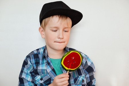 Adorable little boy in trendy shirt and black cap looking at big colorful lollipop. Cute schoolboy wearing fashionable clothes looking sweet candy. People and lifestyle concept.の写真素材