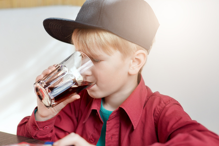 A close-up of adorable little kid boy in stylish cap drinking juice in summer while sitting in restaurant. The boy with a glass of juice isolated over white background. Children and lifestyle.の写真素材