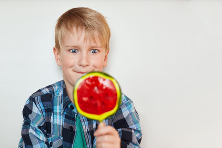 Close up portrait of handsome male child with blond hair and blue eyes dressed in checked shirt holding huge sweet candy in his hand looking with surprise at it wanting to lick itの写真素材