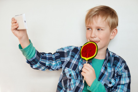 A cute little boy with blond hair and blue eyes dressed in modern shirt holding sweet lollipop in his hand and smartphone making selfie. People, technology and fashion concept.の写真素材